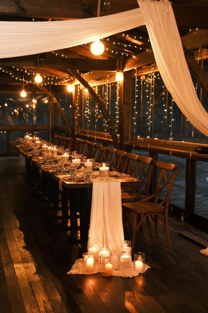 Romantic candlelit dinner setup with draped fabric and twinkle lights in rustic venue.